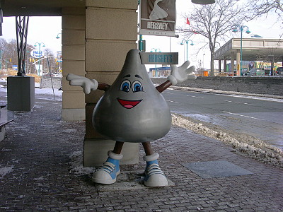 [A statue of a silver Hershey kiss with eyes, eyebrows, an open mouth, and feet with blue sneakers, and arms with white gloves stands near a column of the building.]